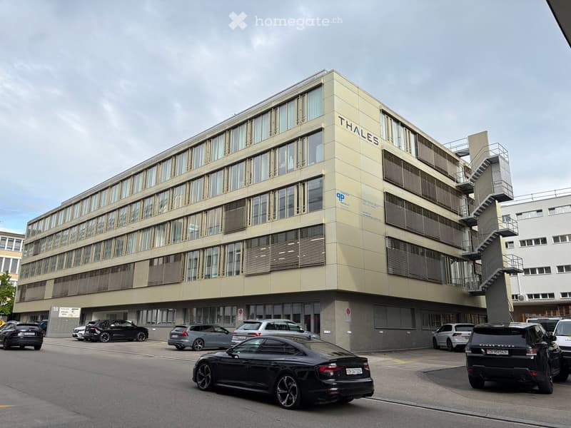 Modern multi-story building with large windows and a sign reading "THALES" on the facade, surrounded by parked cars.