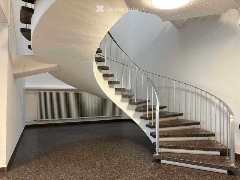 Spiral staircase with dark stone steps and a modern metal railing, leading up to a higher level. The hallway has a dark, textured floor and white walls with a curved ceiling.