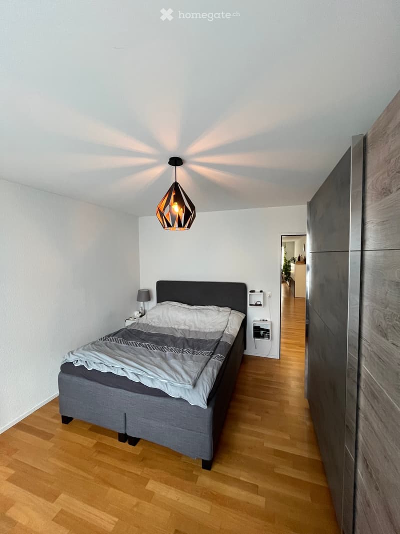 Modern bedroom with a grey upholstered bed, nightstand, and a stylish geometric pendant light fixture. The room features light wooden flooring and white walls.