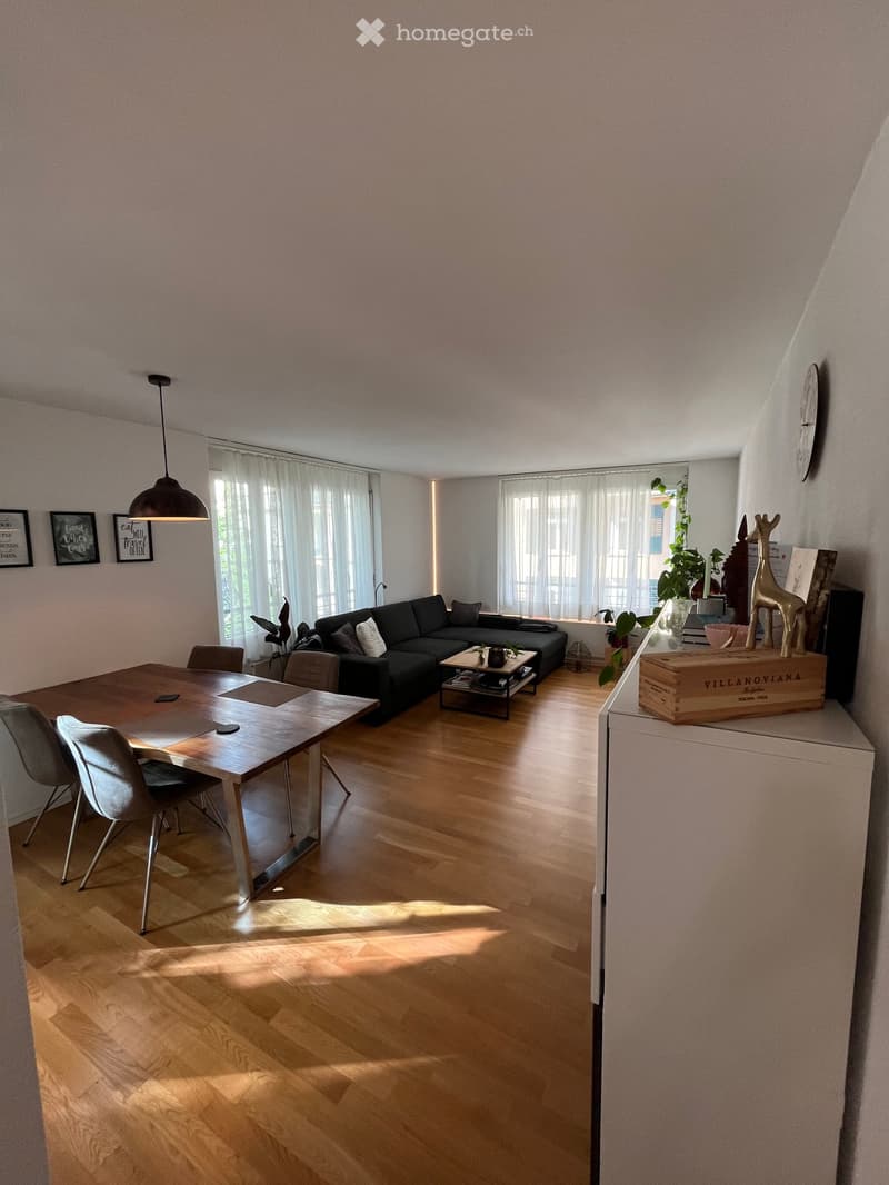 Bright living and dining area with wooden flooring, large windows with white curtains, a black sectional sofa, a wooden coffee table, a dining table with chairs, and a pendant light fixture.