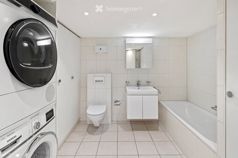 Modern bathroom with white tiles, a toilet, a sink with a mirror, a bathtub, and a stacked washer/dryer unit.