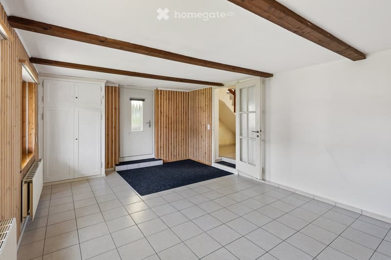 Empty room with white tiled floor, white walls, wooden ceiling beams, white cabinet, and a small black rug. No furniture or equipment present.