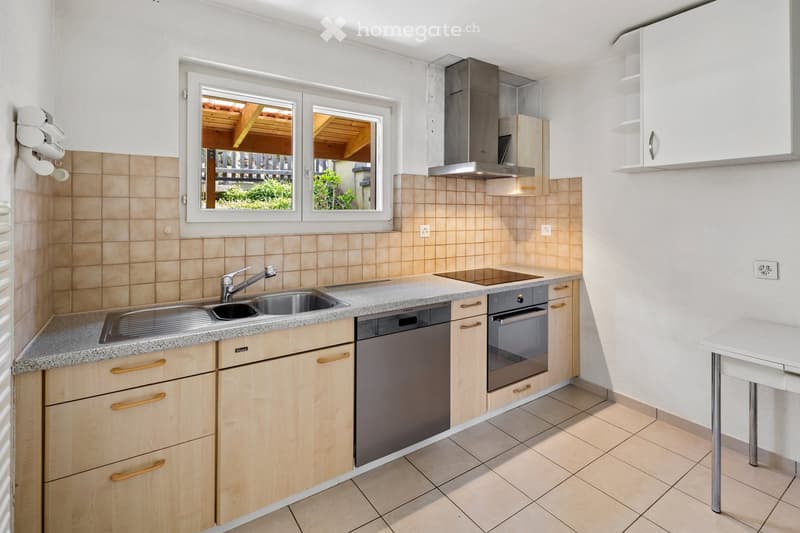 Modern kitchen with light wooden cabinets, granite countertop, stainless steel sink and dishwasher, built-in oven, and a small table near the window.