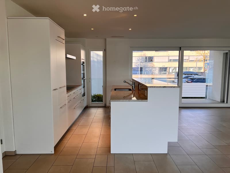 Modern kitchen with white cabinets, a central island, and a sink area. The space features tiled flooring and large windows offering a view of the street.