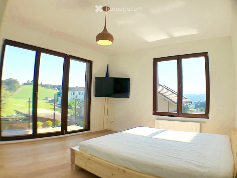 Bright bedroom with large windows and sliding doors, wooden bed, TV mounted on wall, radiator, and modern lighting fixture.