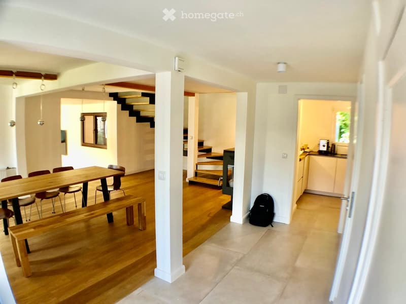 Open-plan living and dining area with wooden flooring, a large wooden dining table with chairs, a bench, a staircase leading to an upper level, and a visible kitchen area with a counter and appliances.