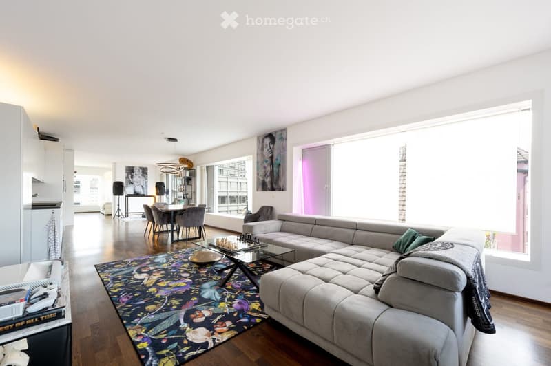 Modern living room with a grey sectional sofa, glass coffee table, colorful rug, dining area with chairs and table, and large windows letting in natural light.