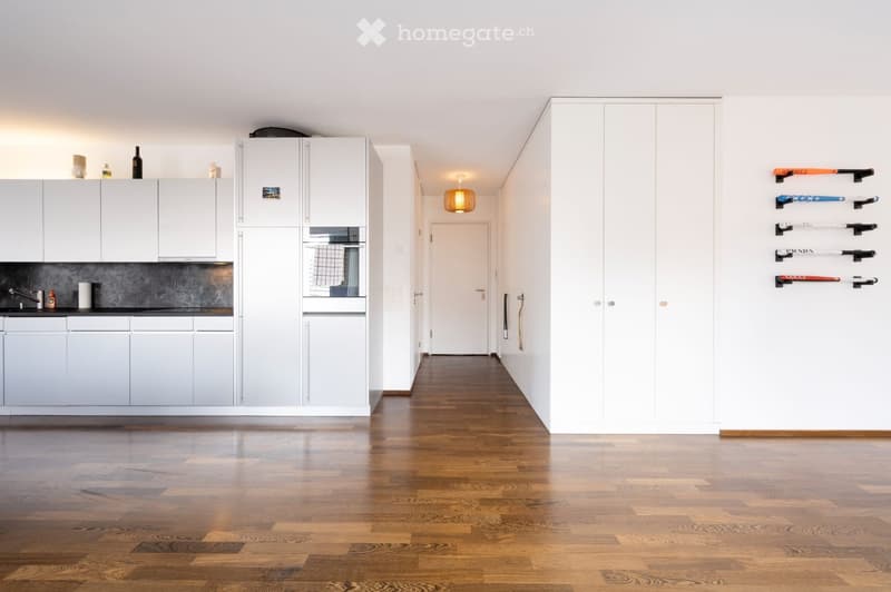 Modern hallway with wooden flooring, white walls, built-in storage units, and decorative skis mounted on the wall. The hallway leads to a kitchen area with white cabinets and a sink.