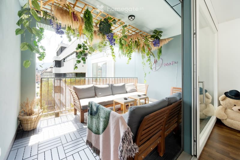 Balcony with a wooden sofa, small table, chairs, and a bench, decorated with hanging plants and a blanket draped over the bench.