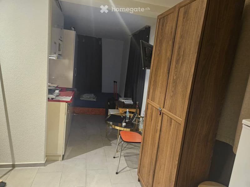 A small, furnished room with a bed, a small table with a laptop, a chair, a refrigerator, and a microwave. The room has tiled flooring and a partially open wooden door.