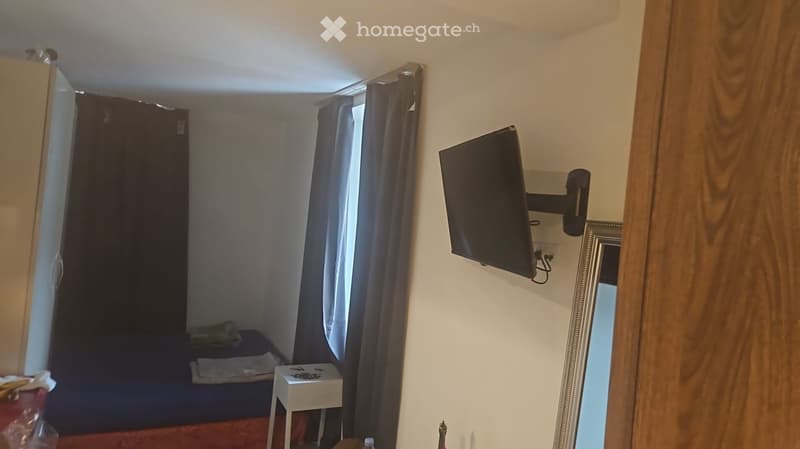 Small bedroom with a bed, nightstand, wall-mounted TV, and a mirror. The room has curtains and a wooden door frame.