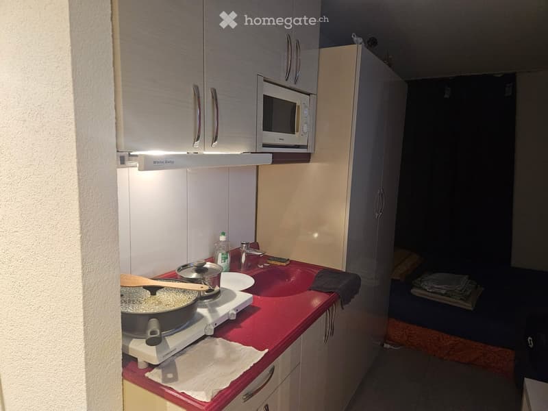 Compact kitchen with white cabinets, red countertop, microwave, stove, sink, and various kitchen items.