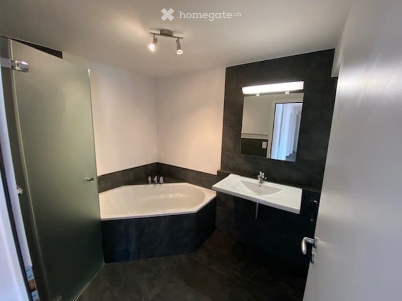 Modern bathroom with a dark grey tiled floor and walls, a white octagonal bathtub, a white sink with a modern faucet, and a large mirror with built-in lighting above the sink.
