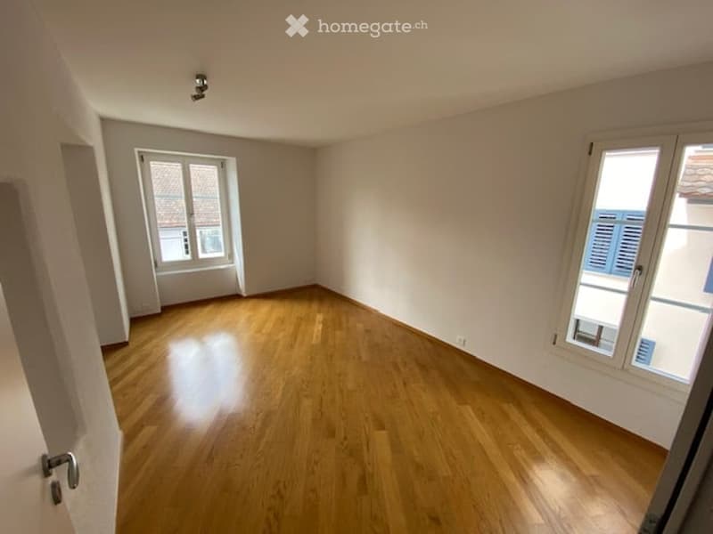 Empty room with wooden flooring, white walls, and two windows with shutters.