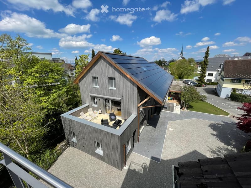 Modern two-story house with grey wooden siding, solar panels on the roof, and a small outdoor seating area on the roof terrace.