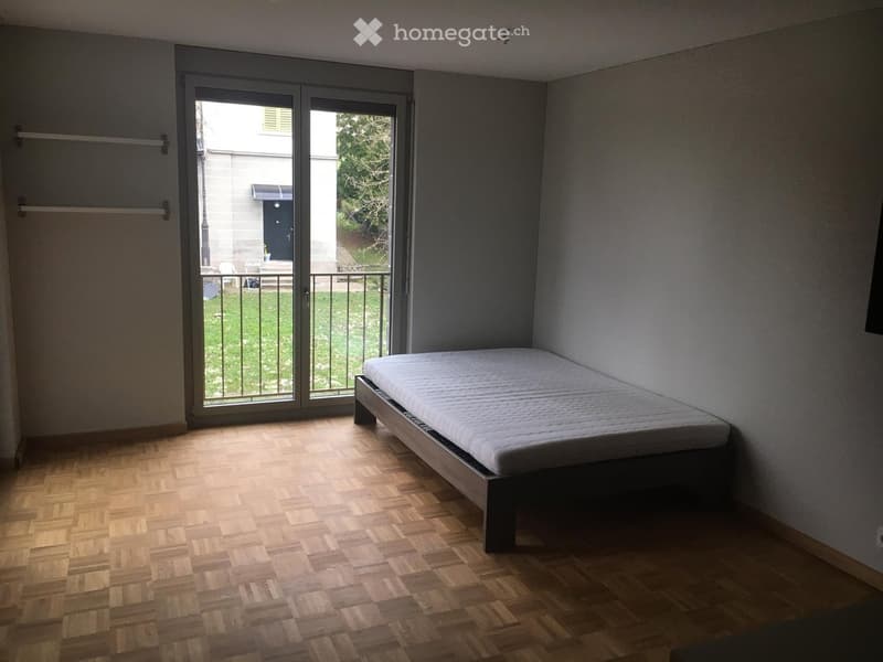 Minimalist bedroom with wooden herringbone flooring, a single bed, and large sliding glass doors leading to a balcony with a view of a garden.