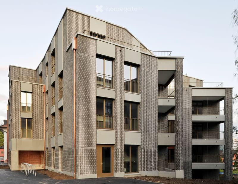 Modern multi-story building with brick facade, large windows, and balconies. Features a central entrance with a wooden door and a small staircase leading to the first floor.