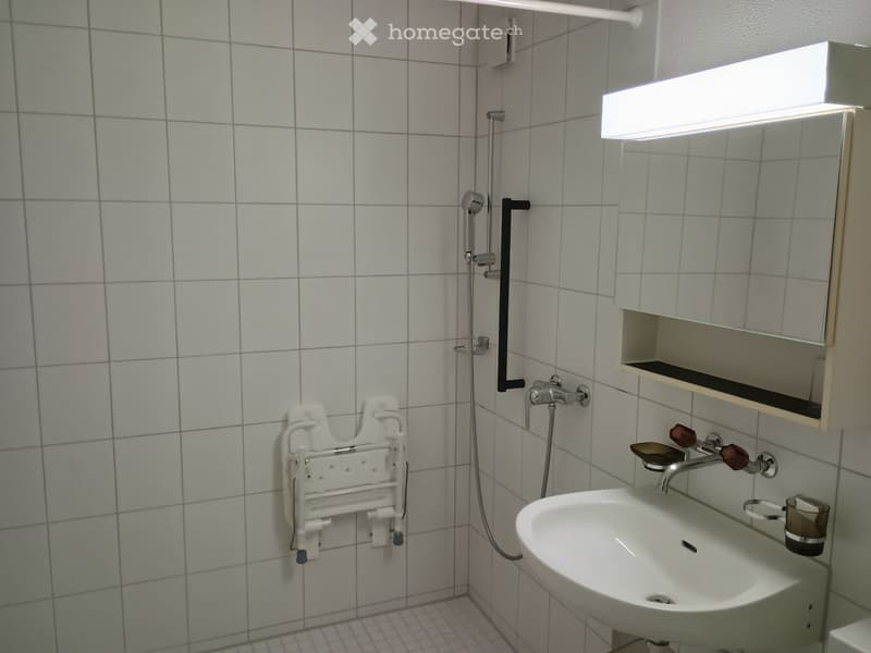 Modern bathroom with white tiled walls and floor, a sink with a modern faucet, a shower head with a rail, a mirror with a built-in shelf, and a cup on the sink.