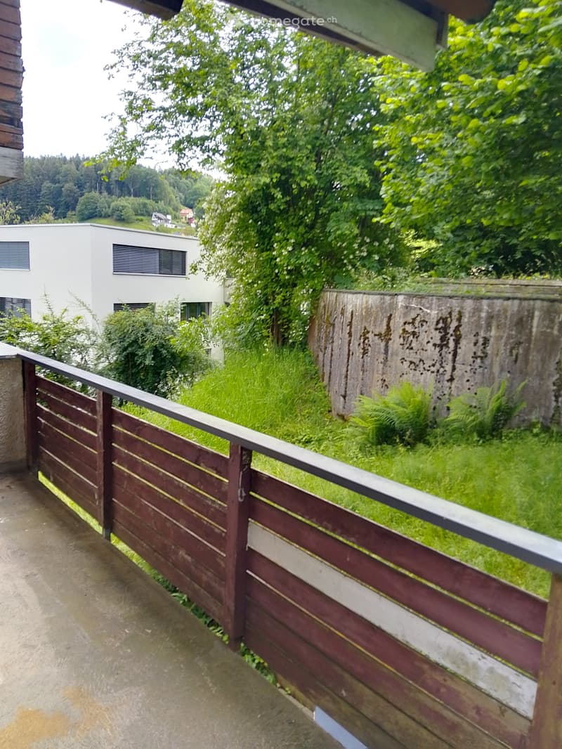 Balcony with wooden railing, lush greenery, and a view of a modern white house in the background.