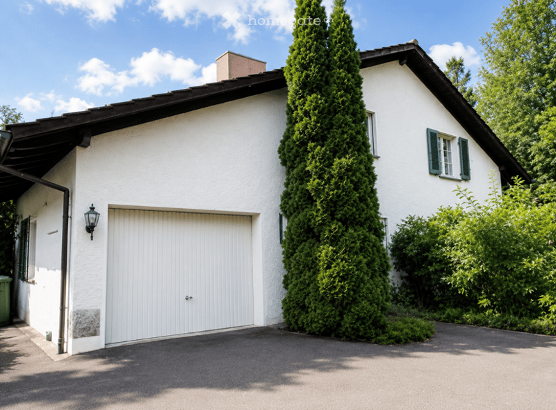 Single-story white house with a garage door, green shutters, and a tall conifer tree in front.