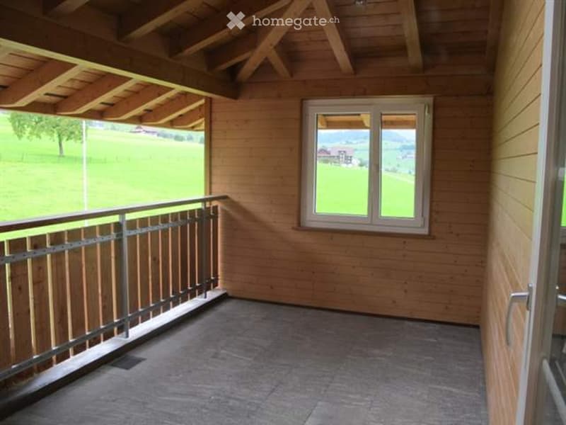 Wooden balcony with metal railing, large window offering a scenic view of green fields and mountains.