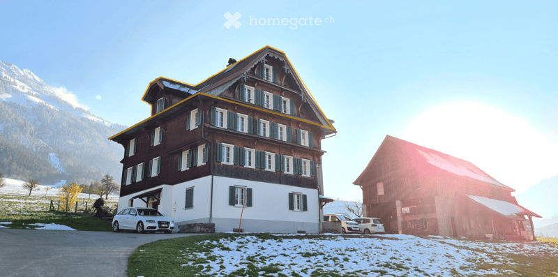Traditional wooden house with white walls, green shutters, and a yellow roof, situated in a snowy landscape with mountains in the background.