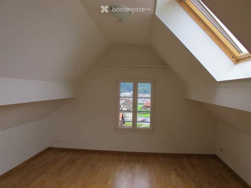 Empty attic room with wooden flooring, white walls, and two windows offering a view of the outside. The room features a sloped ceiling with a skylight.