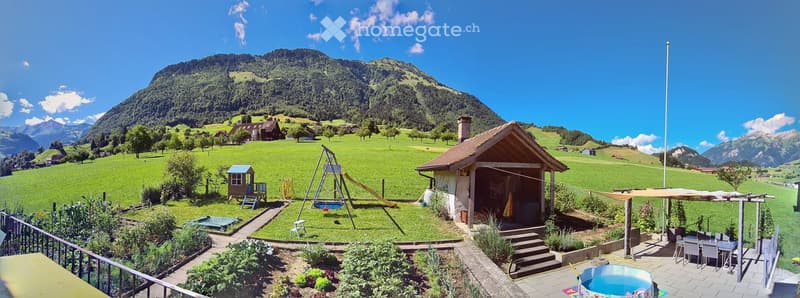 A picturesque garden area with a small wooden cabin, a swing set, a slide, a patio with a dining table and chairs, and a small swimming pool, all set against a backdrop of green hills and mountains.