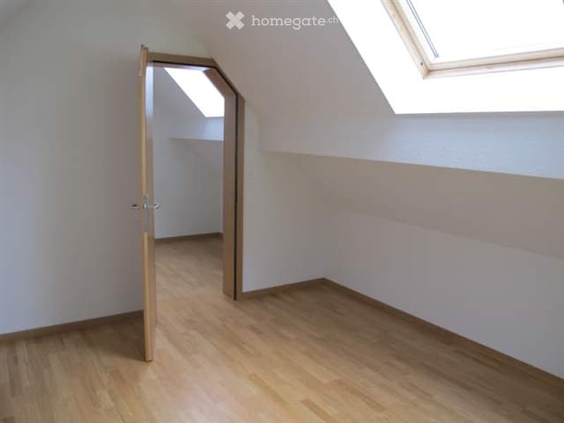 Empty room with wooden flooring, white walls, and a skylight window. The door is open, revealing another room with similar features.