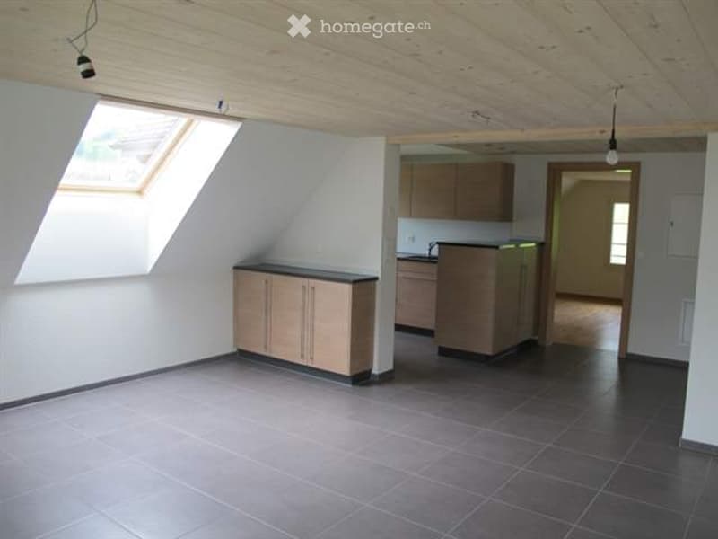 Bright, unfurnished room with exposed wooden beams, tiled floor, and built-in cabinets. A small kitchen area is visible with a sink and cabinets.