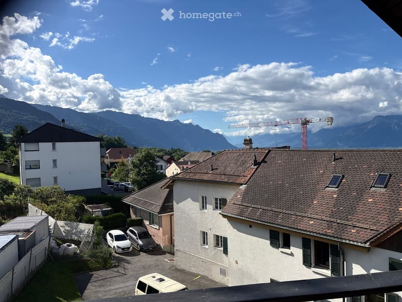 View from a balcony showing a residential area with houses, parked cars, and a mountainous backdrop under a partly cloudy sky.
