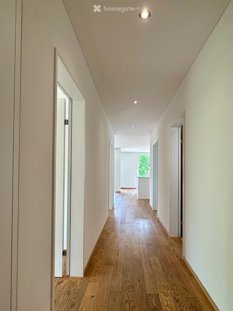 A long, narrow hallway with light wooden flooring and white walls, featuring recessed ceiling lights and several open doors leading to other rooms.