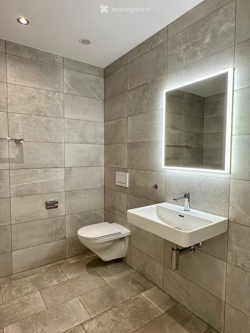 Modern bathroom with grey tiled walls and floor, white toilet, white sink with chrome faucet, and illuminated mirror above the sink.