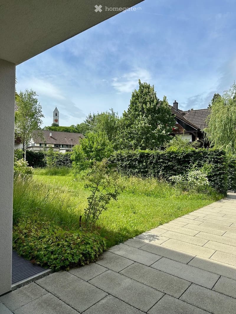 Garden area with a paved pathway, lush greenery, and a view of a town with a church tower in the background.