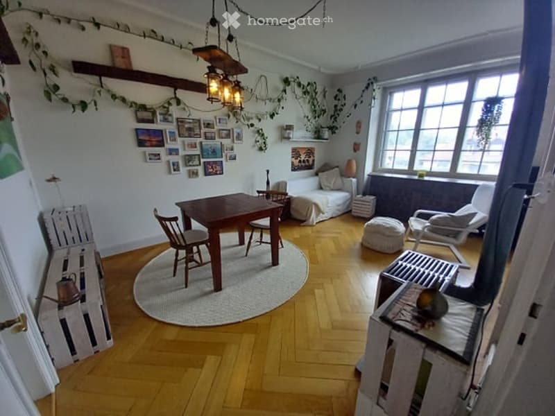 Furnished Room. Open-plan living and dining area with wooden flooring, large windows, a dining table with chairs, a sofa, an ottoman, and decorative picture frames on the wall.