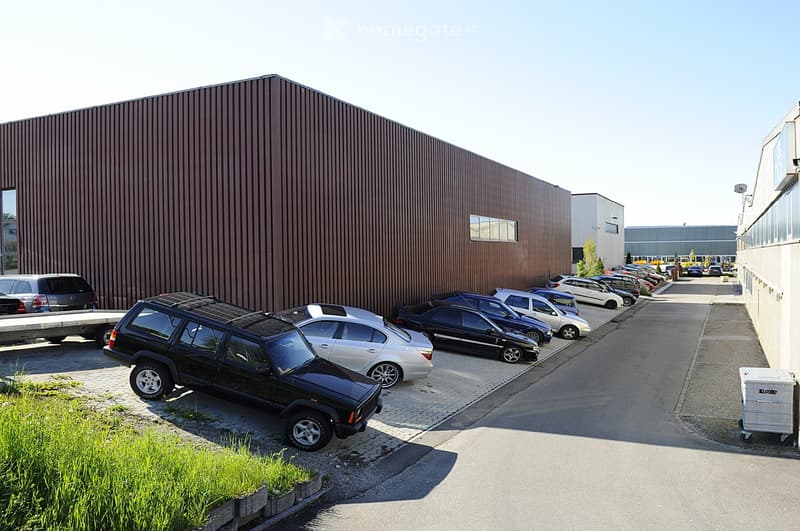 A large industrial-style building with a brown metal facade and a flat roof, surrounded by a parking lot with several cars.