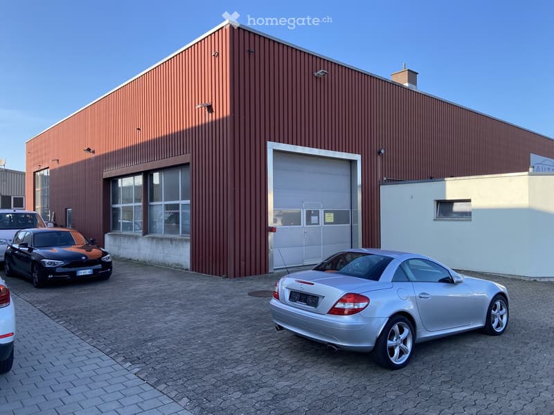 A large industrial-style building with a red metal facade, large garage door, and several windows. Several cars are parked in front of the building on a paved area.