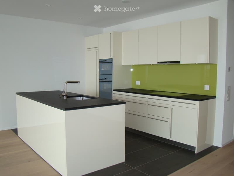 Modern kitchen with white cabinets, black countertops, a sink with a faucet, and an oven. The backsplash is green, and the floor is dark tiled with a wooden border.