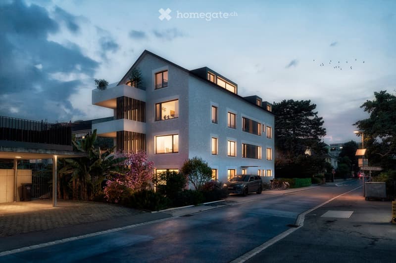 Modern multi-story residential building with large windows, illuminated at dusk, surrounded by greenery and a paved driveway.