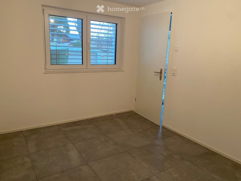 Empty room with tiled floor, white walls, and a window with blinds. A white door is slightly open, revealing a glimpse of the outside area.