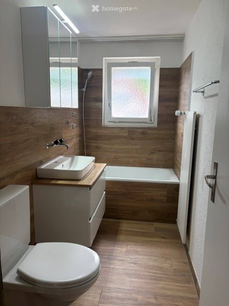 Modern bathroom with wooden walls and floor, white fixtures including a sink, toilet, and bathtub, a large window, and a built-in radiator.