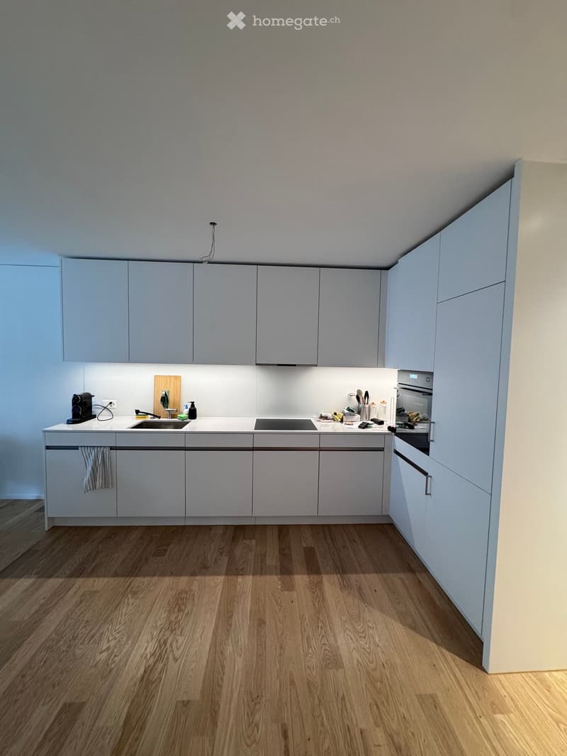 Modern kitchen with light grey cabinets, white countertops, and wooden flooring. Features a sink, faucet, oven, coffee maker, cutting board, and a towel hanging from a cabinet.