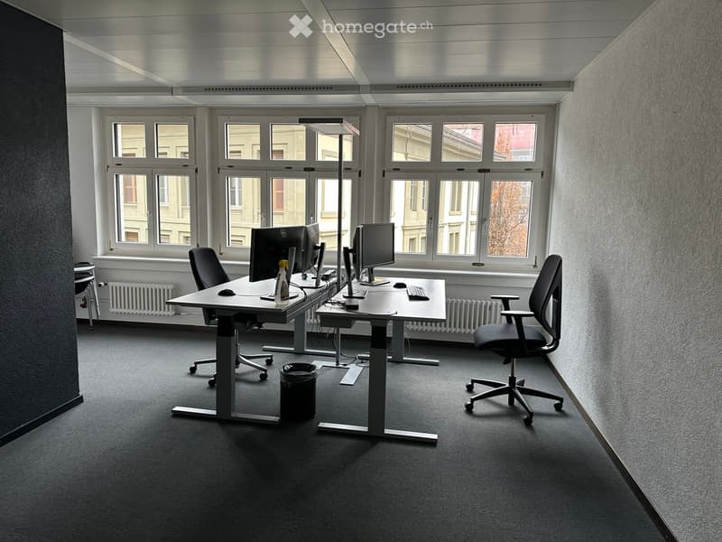 Modern office space with two desks, ergonomic chairs, computer monitors, keyboards, and mice. Large windows provide natural light, and the room features a carpeted floor and radiators.
