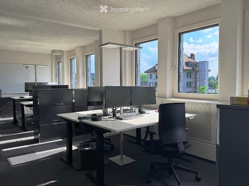 Modern office space with desks, chairs, computer monitors, keyboards, and mice. Large windows provide natural light, and the room is equipped with lamps for additional illumination.