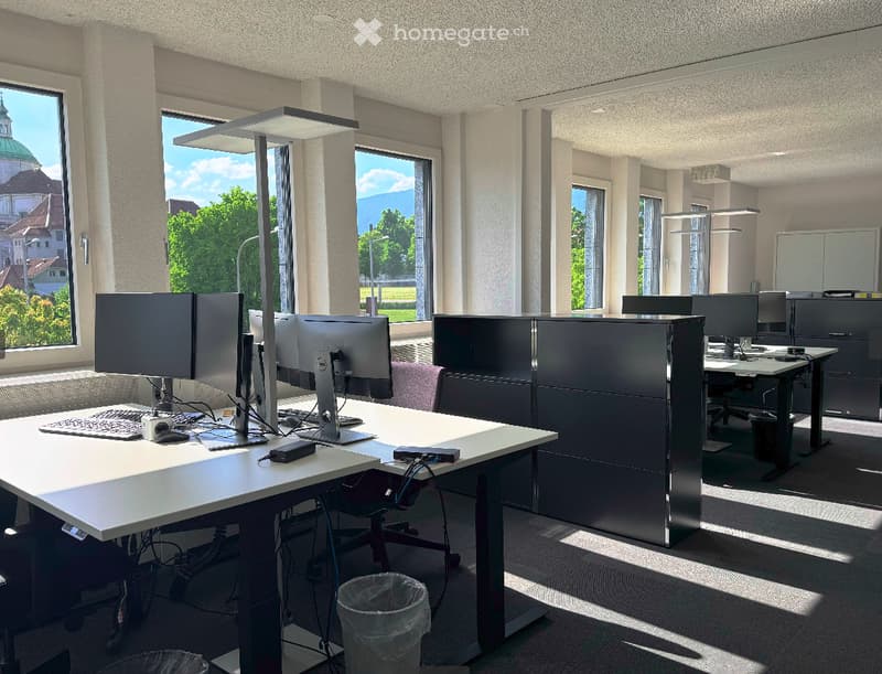 Modern office space with large windows offering a scenic view, equipped with desks, chairs, computer monitors, keyboards, and lamps.