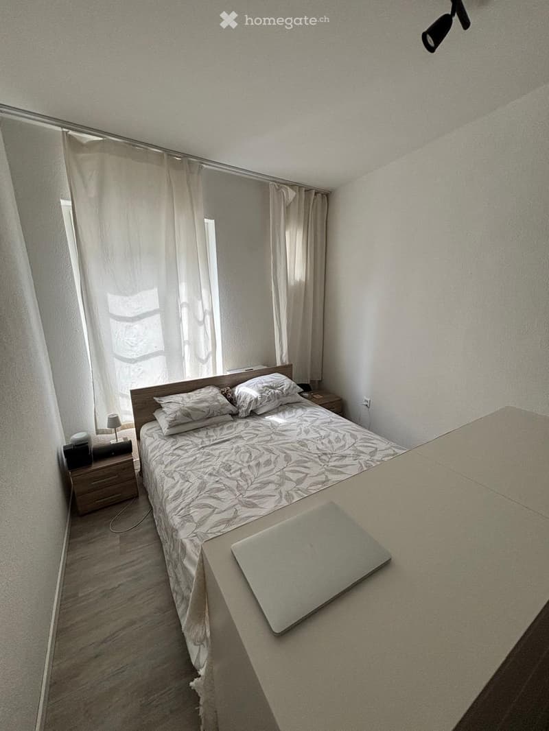 A modern bedroom with a neatly made bed, a laptop on a desk, and a nightstand with a lamp. The room features light-colored walls, wooden flooring, and sheer curtains on the windows.