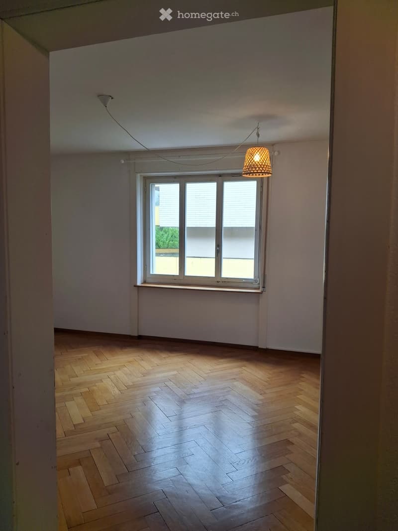 Empty room with wooden herringbone flooring, white walls, and a large window with blinds. A single hanging light fixture is visible.