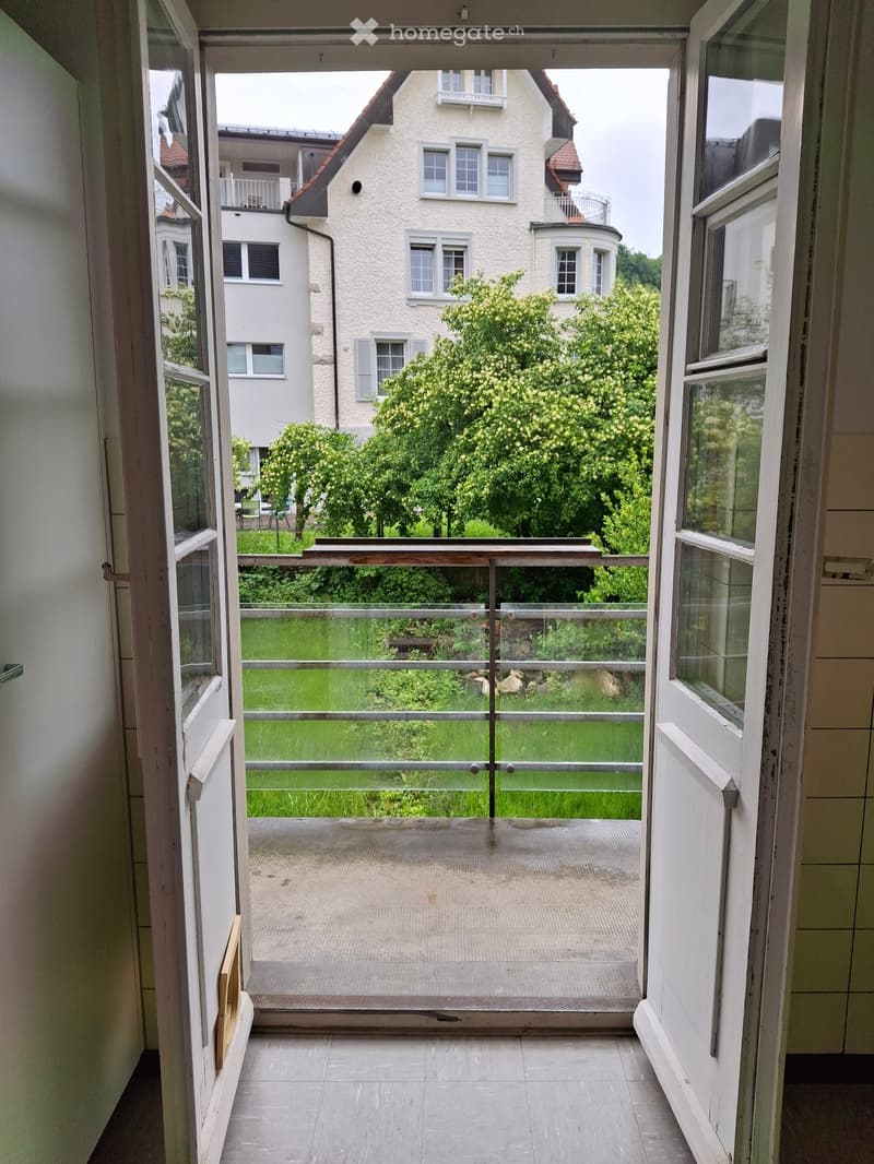 Balcony with glass railing, tiled floor, overlooking a green lawn and neighboring buildings.