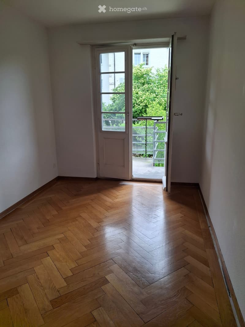 Empty room with wooden herringbone flooring, white walls, and an open sliding door leading to a balcony with a metal railing and outdoor view.