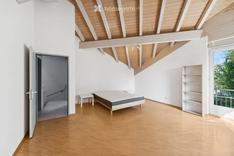 Bright bedroom with wooden flooring, white walls, and a slanted wooden ceiling. Features a bed, a small nightstand, and a white shelving unit near a large window with a view.